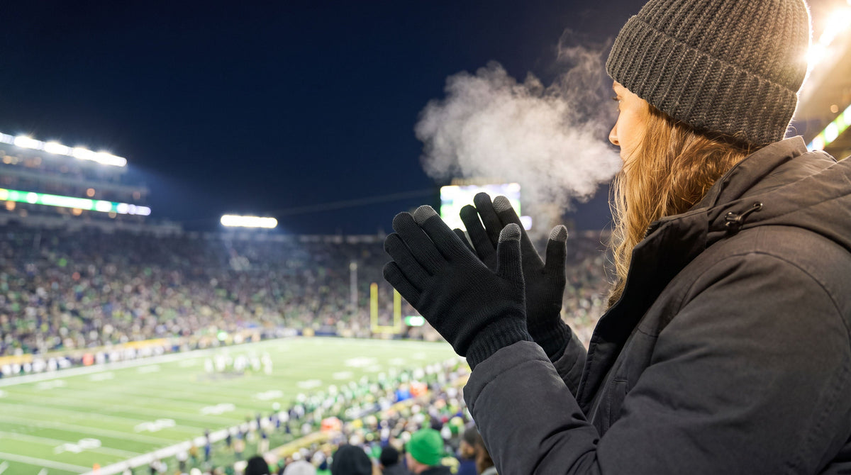 Football fan clapping in the stands at a cold night game, wearing black FibreHeat merino gloves and a knit hat