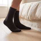 Feet in charcoal FibreHeat Thermal Therapy Socks on an oak floor beside a bed