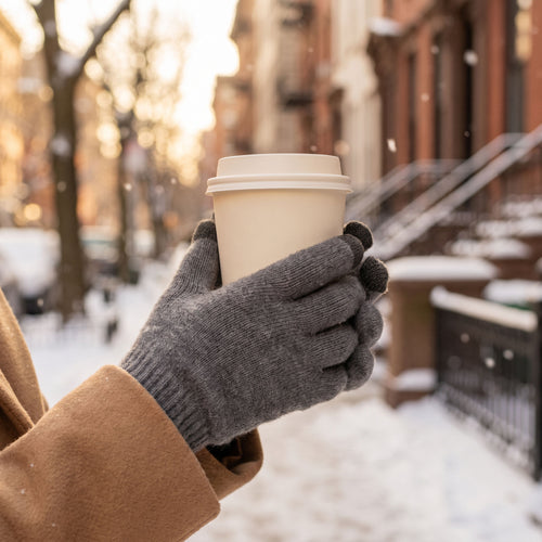 Hands in gray heather FibreHeat cashmere gloves holding a takeaway coffee on a snowy city street