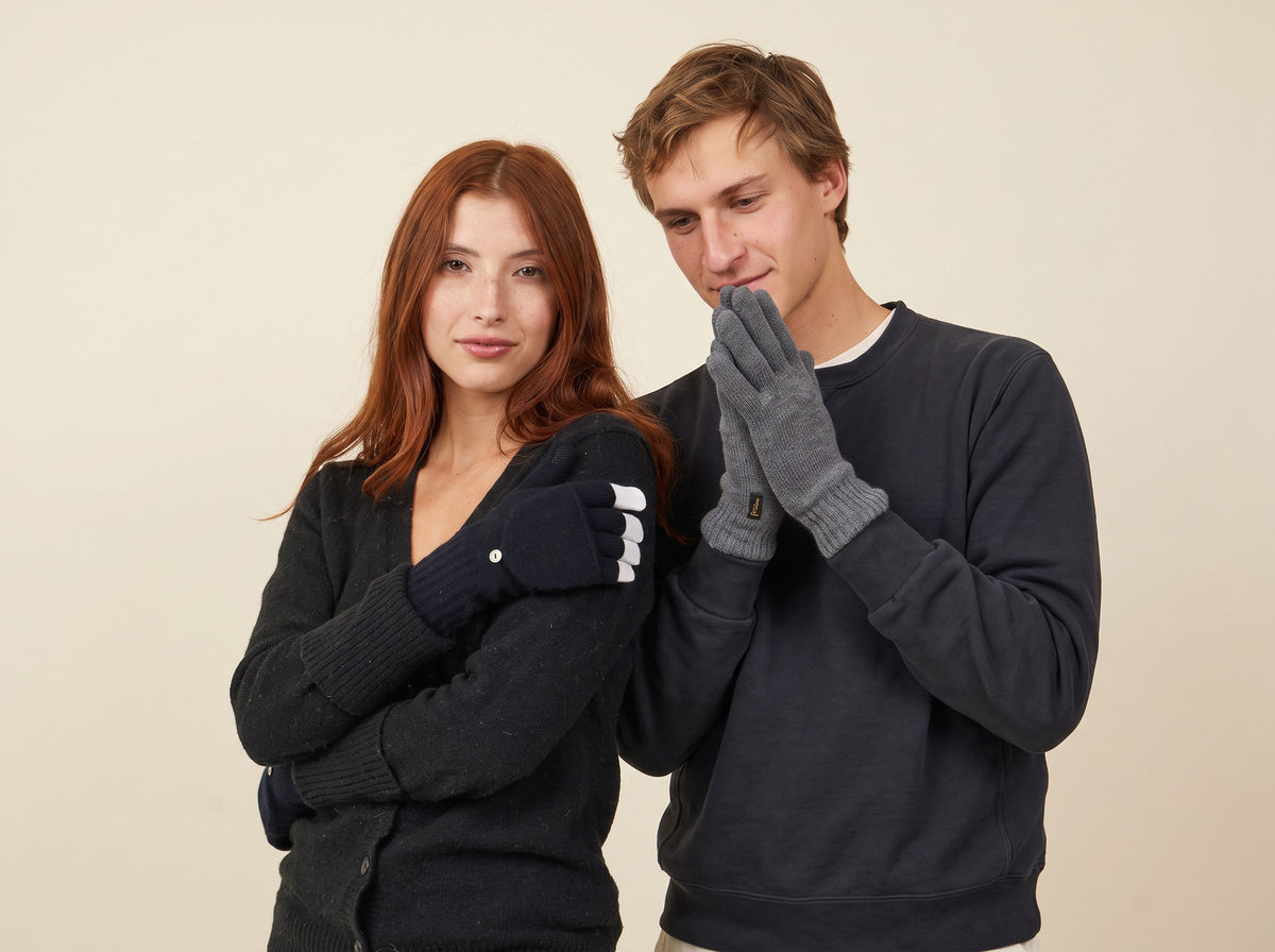 A red-haired woman in black flip-top mittens and a young man in gray merino gloves standing together against a cream backdrop