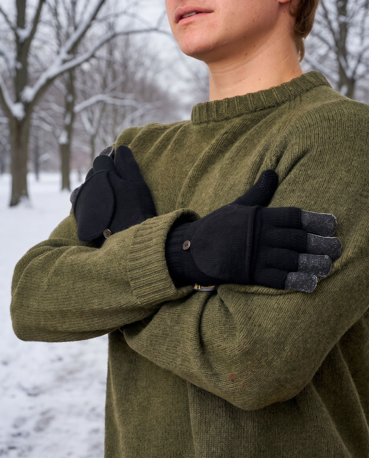 Man in an olive sweater wearing black FibreHeat flip top mittens, one top folded back, in a snowy park