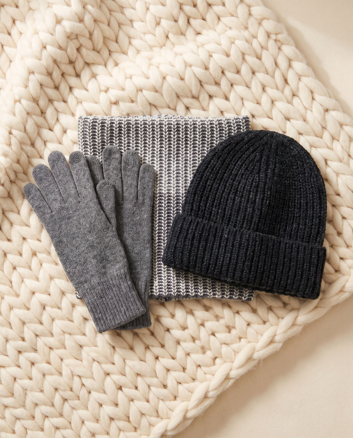 The Head-to-Hand Heat Bundle: cashmere gloves, gray and ecru neck warmer and black knit beanie on a cream knit throw